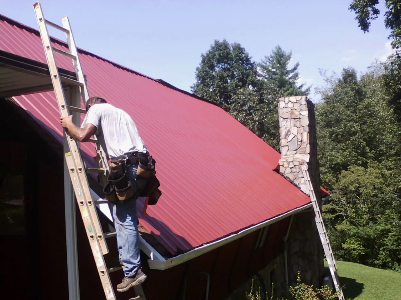 Roofer on a ladder beside a red metal roof and stone chimney — Soffit & Fascia Repair by Roofing Brothers in Scottsboro